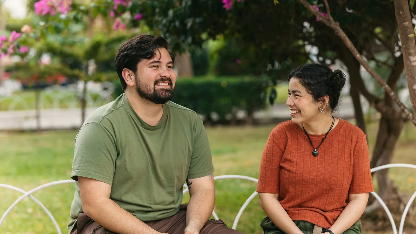 Two people sitting together on a bench in a sunlit garden, laughing in conversation.