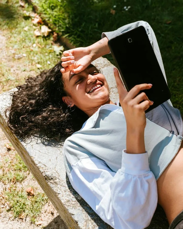 A woman lying on a bench in a sunlit garden, smiling while reading on her tablet.