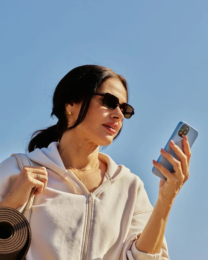 A woman with a yoga mat checks her phone in bright daylight.